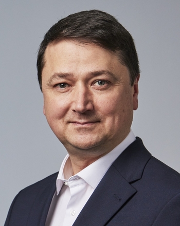 A portrait of a man standing in front of a grey wall, smiling at the camera.
