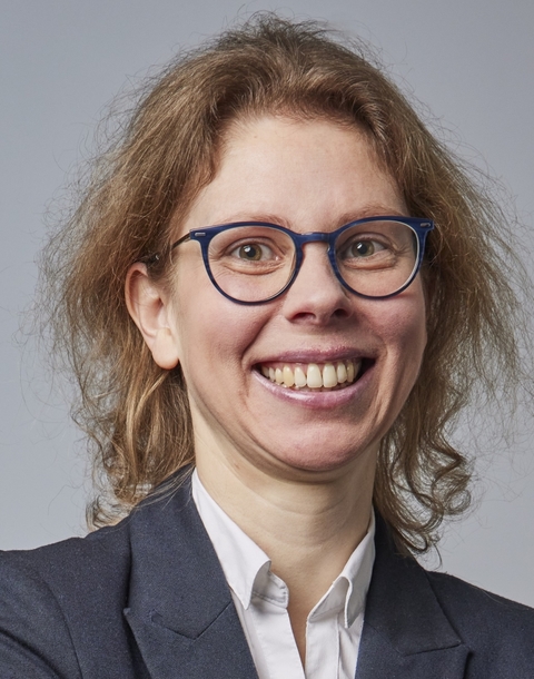 A portrait of a woman standing in front of a grey wall, smiling at the camera.