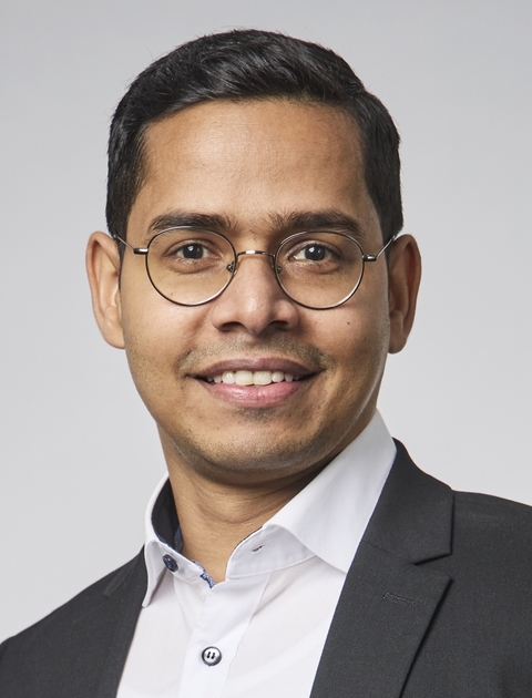 A portrait of a man standing in front of a grey wall, smiling at the camera.