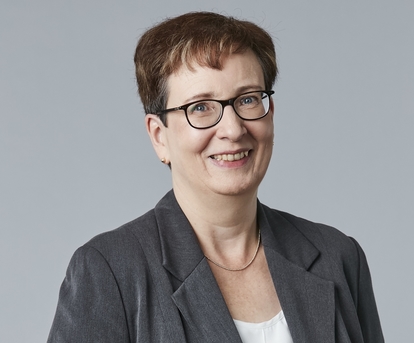 A portrait of a woman standing in front of a grey wall, smiling at the camera.